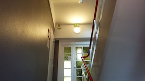 A narrow hallway with a door at the end, a red handrail, and a ceiling light. - ABC Hôtel (Toul)