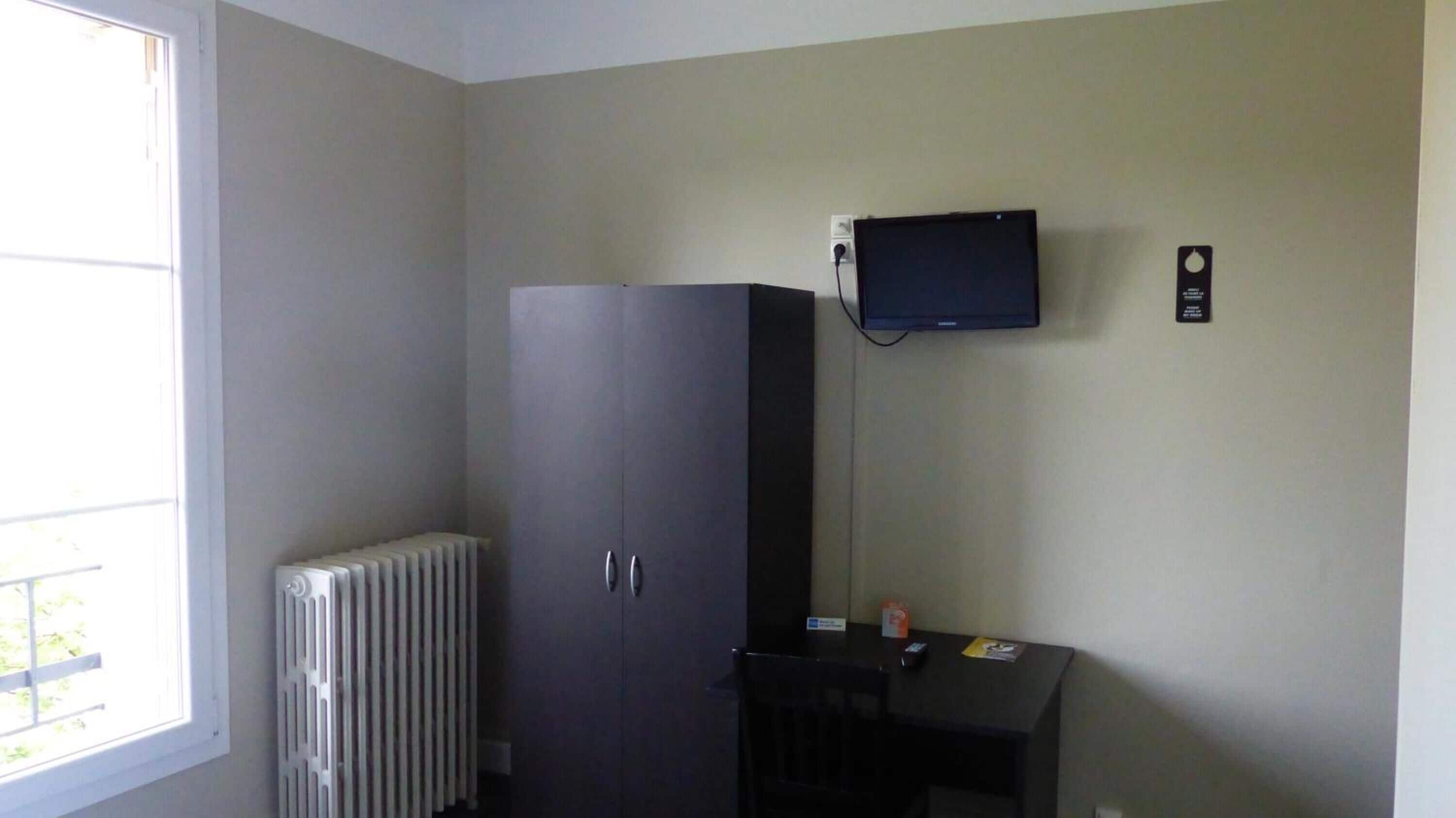 Ein Zimmer mit Heizkörper, Kleiderschrank, einem kleinen Tisch und einem an der Wand befestigten Fernseher sowie einem Fenster.