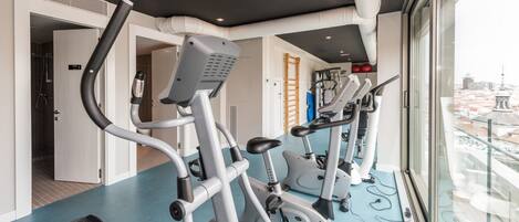 Et fitnesscenter med træningsudstyr, et blåt gulv og udsigt til byen gennem store vinduer.