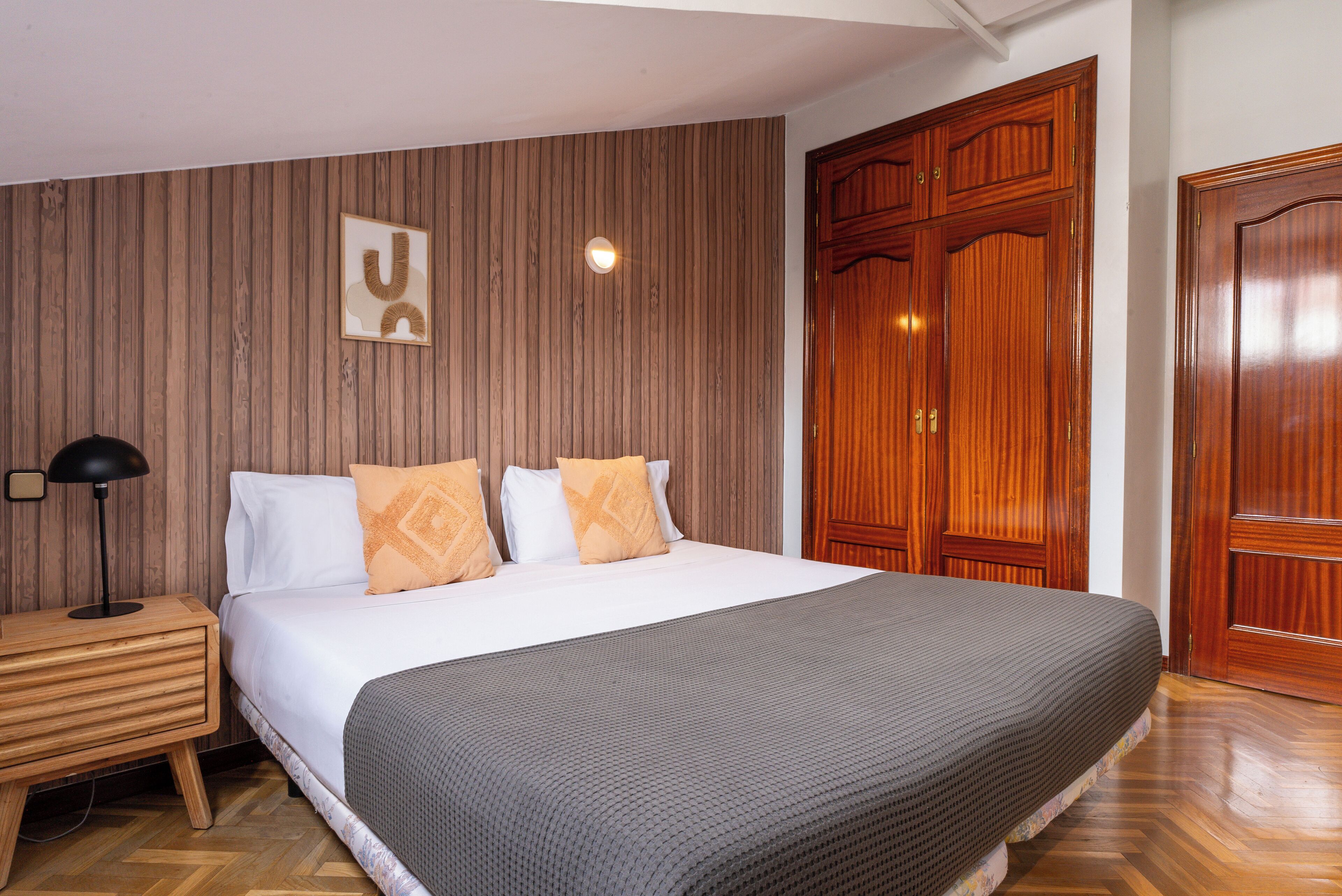 Un dormitorio con cabecera de madera, cama con almohadas blancas y anaranjadas, mesita de noche con lámpara y un armario grande.