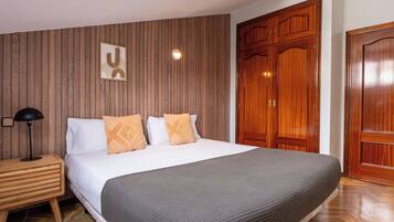 Un dormitorio con cabecera de madera, cama con almohadas blancas y anaranjadas, mesita de noche con lámpara y un armario grande.
