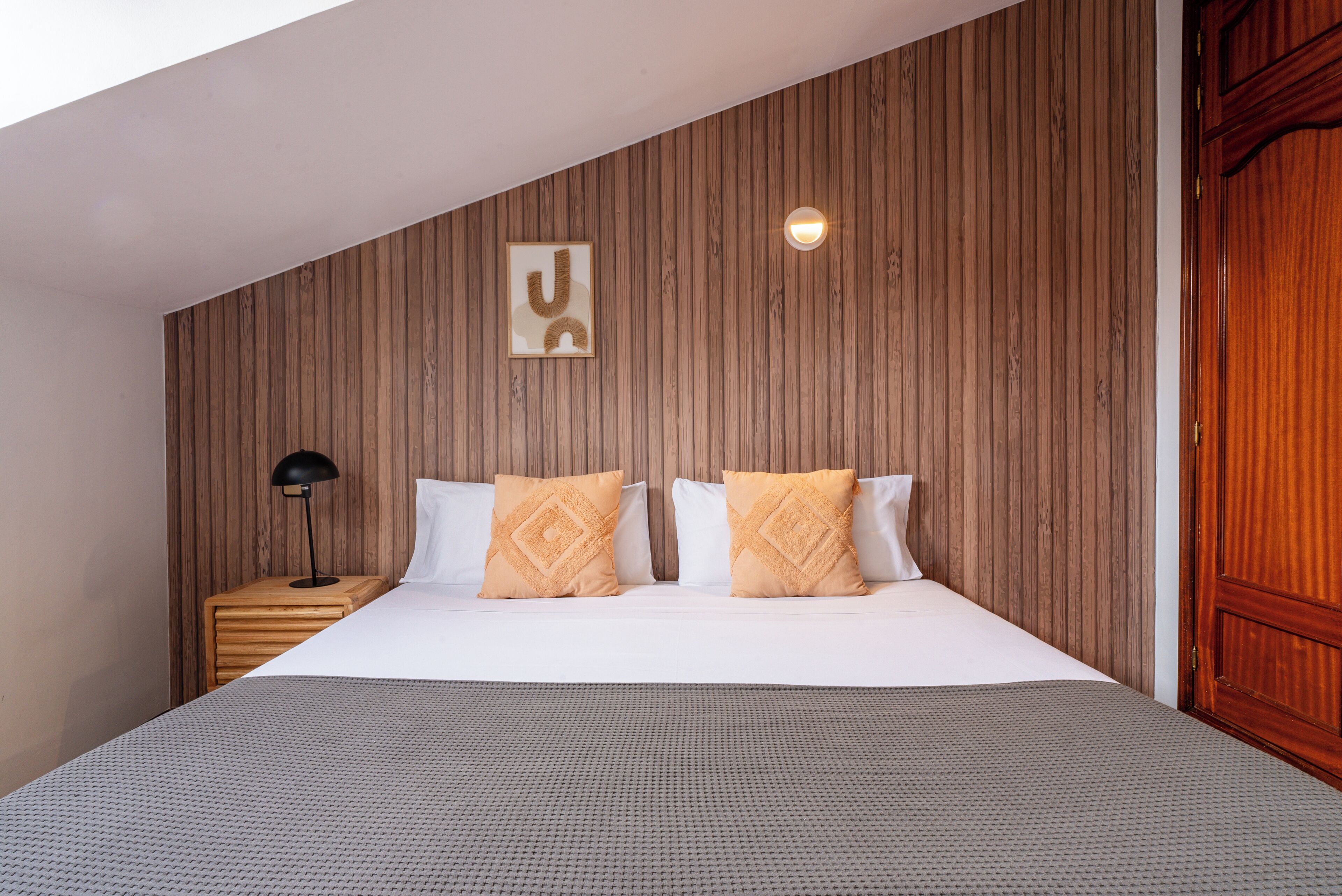 Une chambre avec un plafond en pente, un lambris en bois, un lit avec des oreillers blancs et à motifs, une table de chevet avec une lampe et une porte en bois.