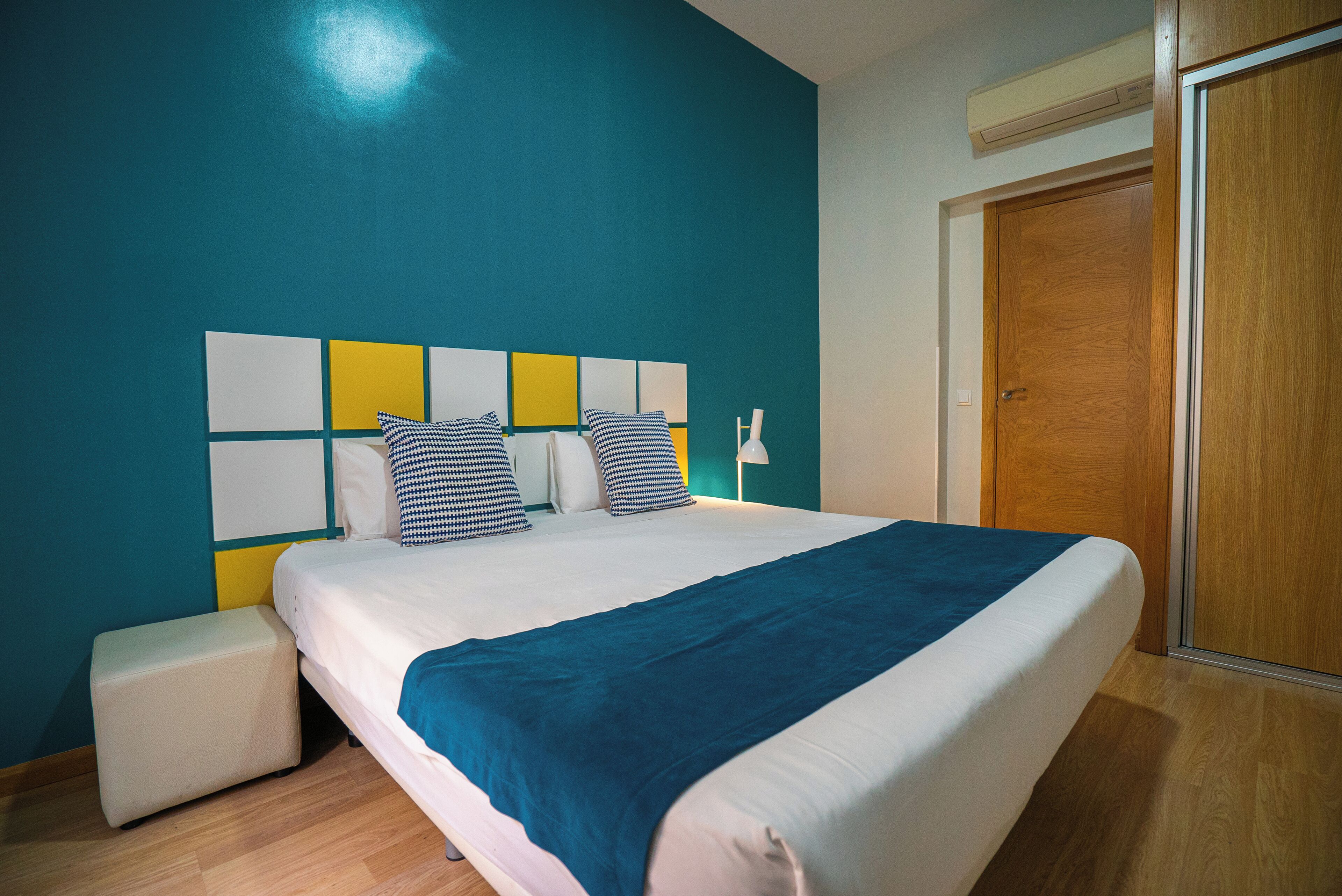 A neatly made bed with a blue blanket, a bedside table, and a wooden wardrobe in a room with teal walls.