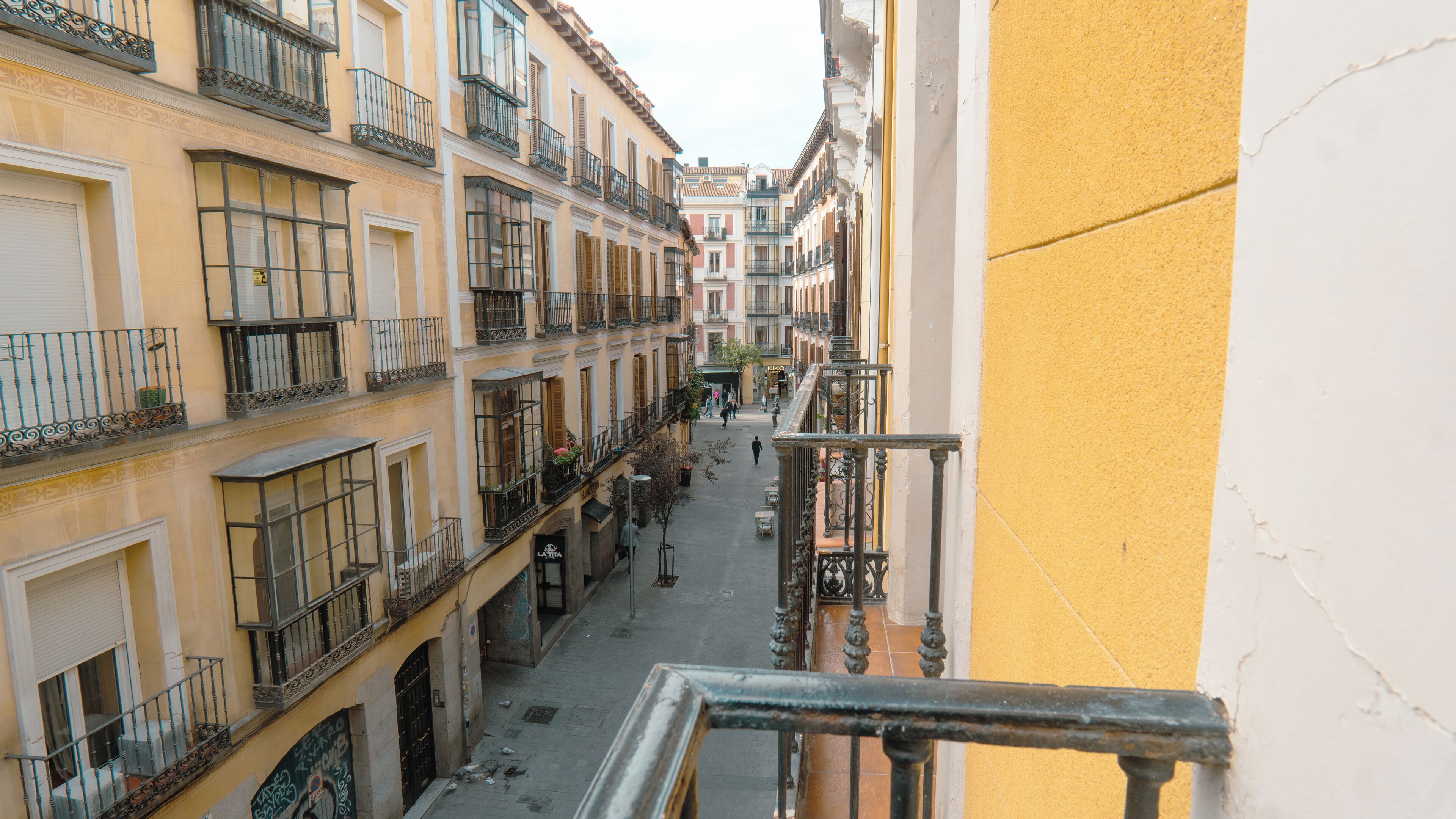 Una calle estrecha flanqueada por edificios con balcones, un escaparate visible y un cielo despejado.