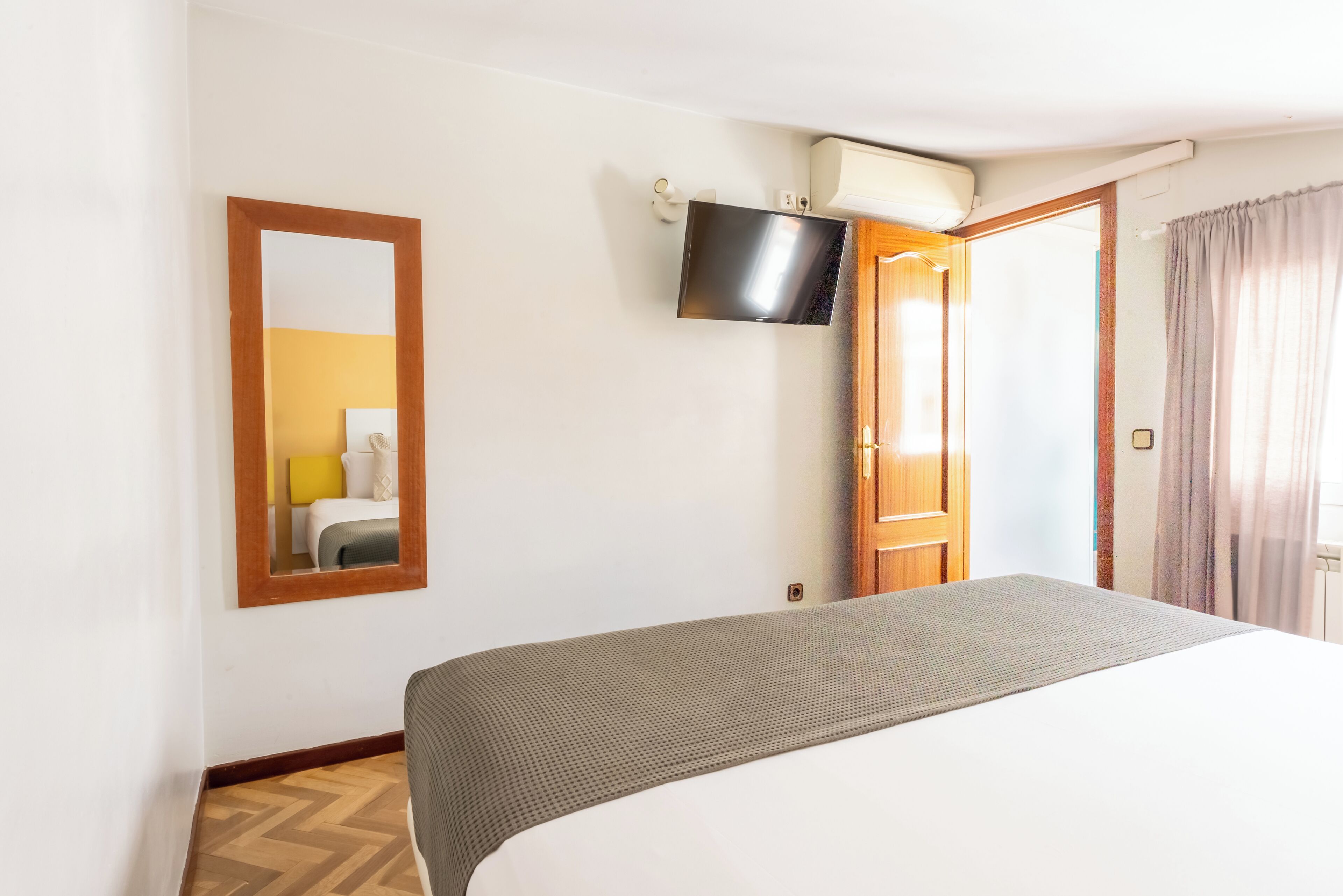 A hotel room with a bed, a TV mounted on the wall, a wooden door, and a mirror.