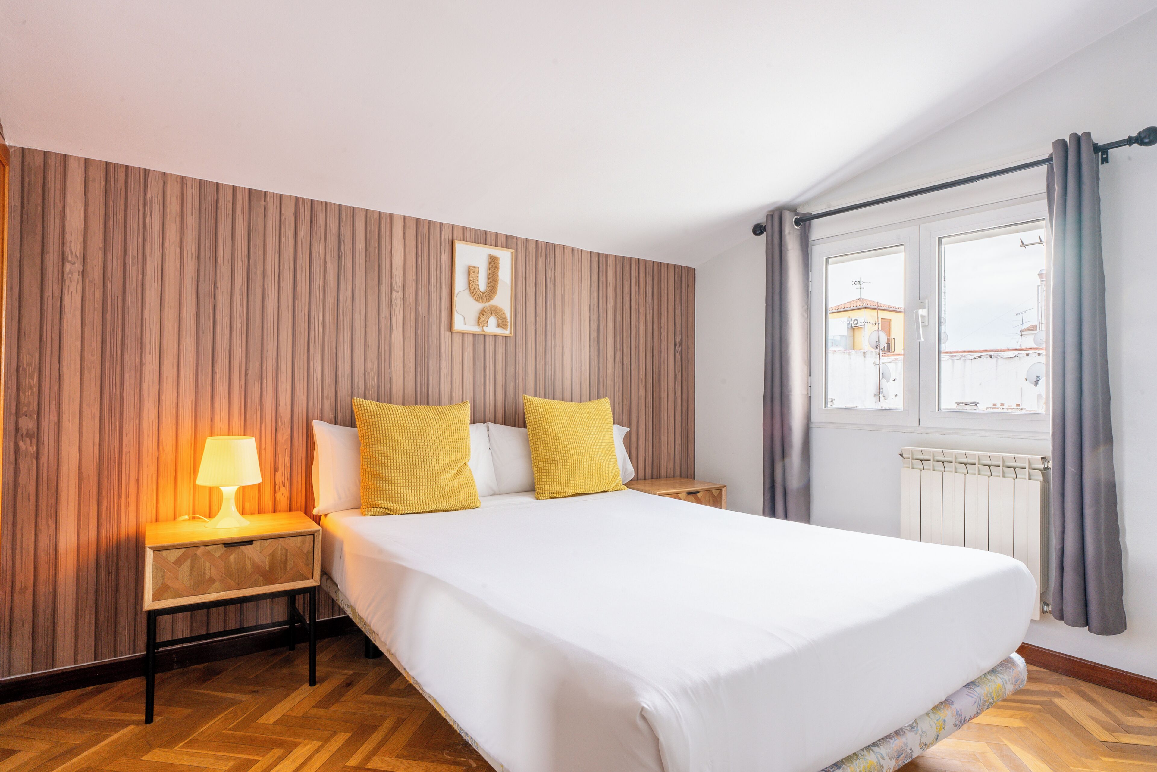 Une chambre d’hôtel avec un mur lambrissé en bois, un lit avec une literie blanche et deux oreillers jaunes, une table de chevet avec une lampe et une fenêtre avec des rideaux.