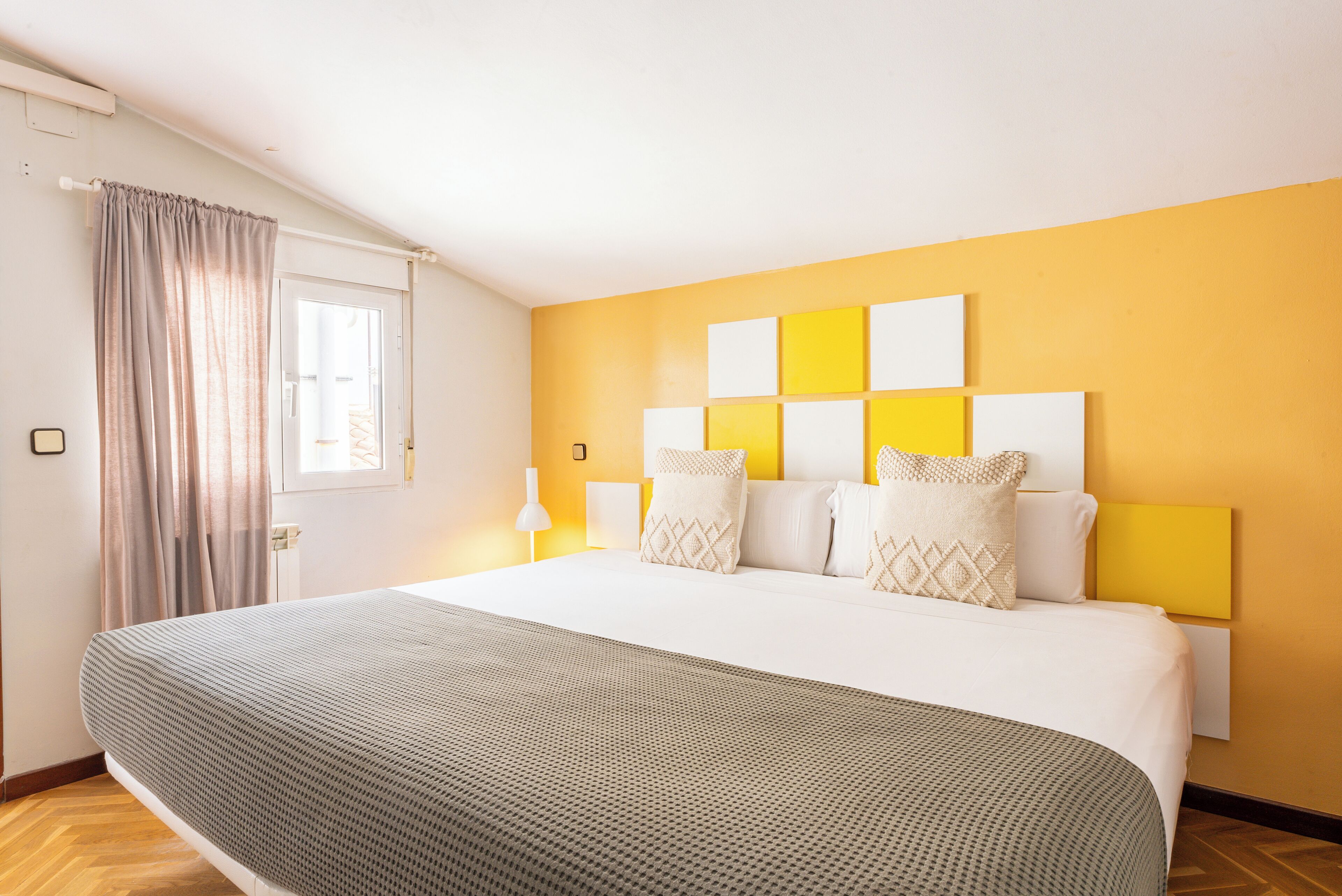 A hotel room with a large bed, a window with curtains, a headboard with multiple pillows, and a yellow accent wall.
