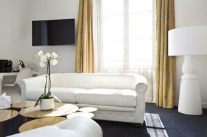 Una habitación de hotel moderna con un sofá blanco, un televisor de pantalla plana, una cafetera, una mesita con una planta en maceta y una lámpara.