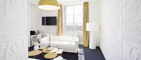 A modern hotel room with a white sofa, a coffee table, a lamp, and a TV mounted on the wall.