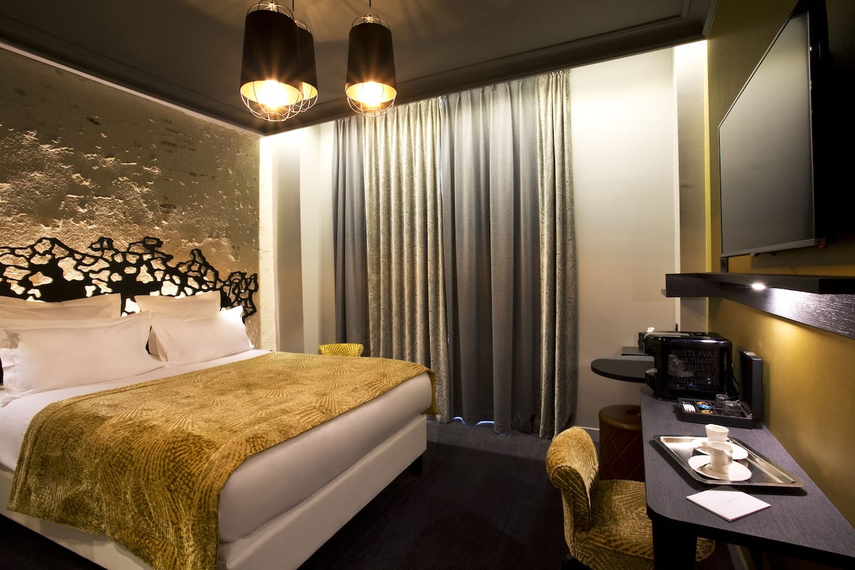 A hotel room with a bed, a desk with a coffee maker, a chair, and a large window with curtains.