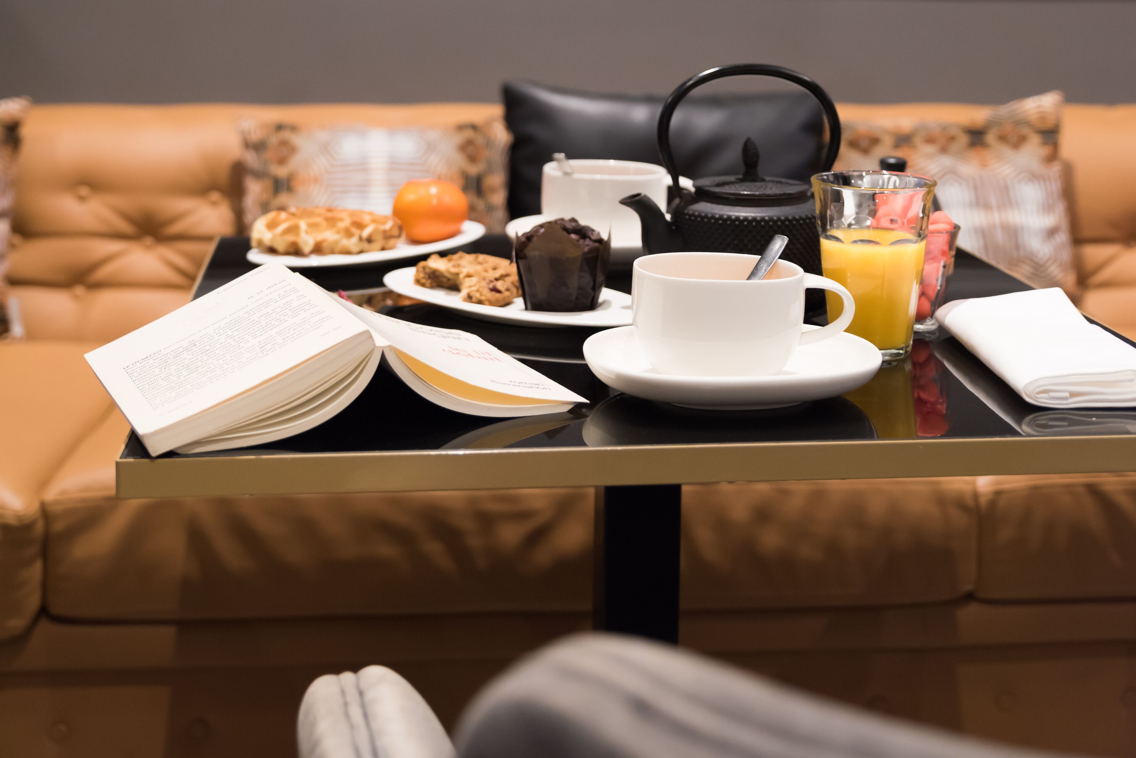 Desayuno con un libro, café, té, jugo de naranja y pasteles sobre una mesa en una habitación con un sofá de cuero marrón.