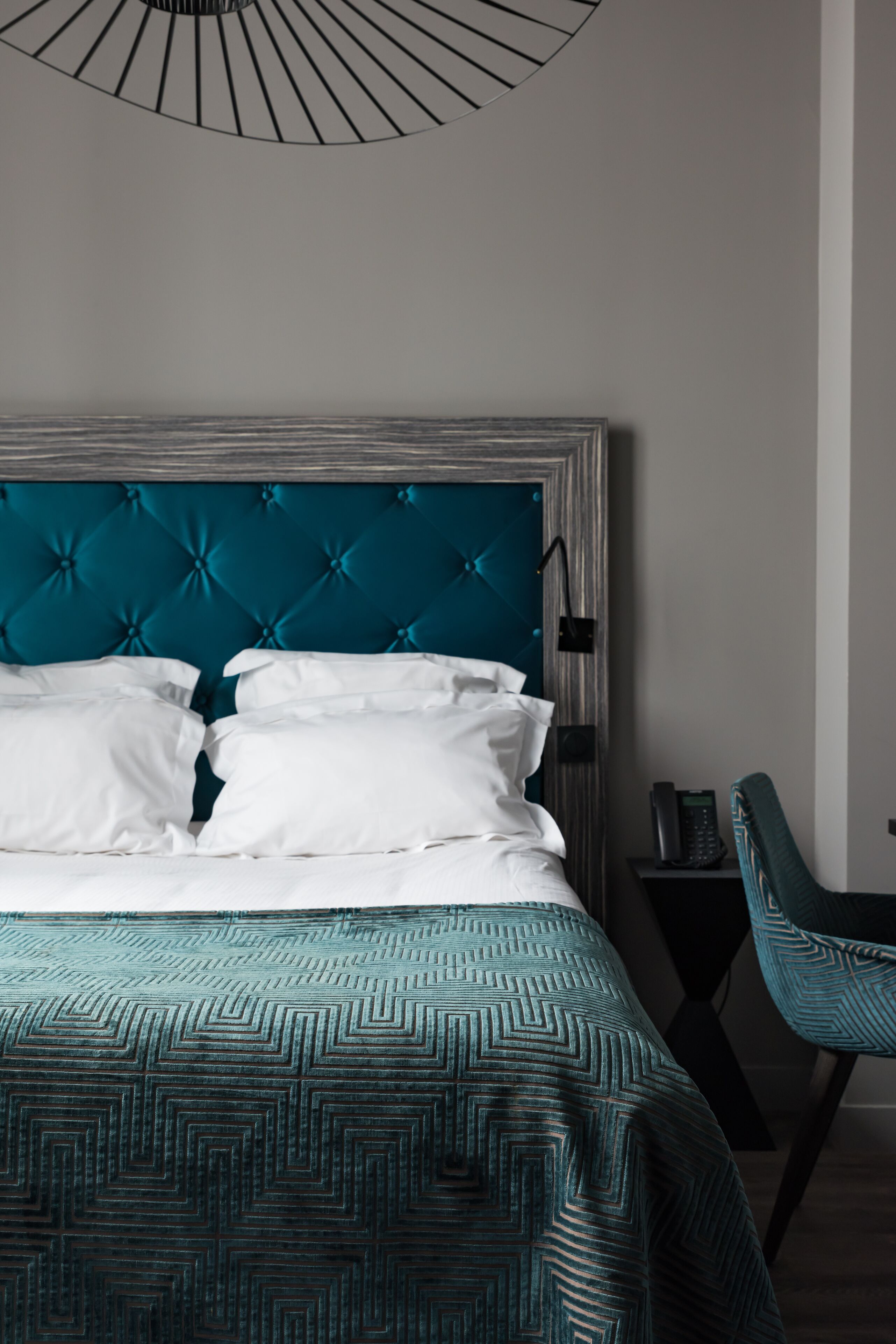 A neatly made bed with white linens and a teal headboard, a matching teal chair, and a modern wall-mounted light fixture.