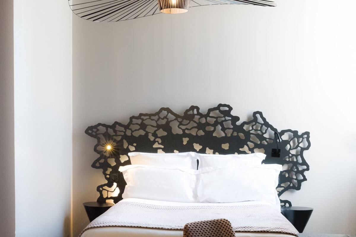 A neatly arranged bedroom with a modern bed, a unique headboard, a woven chair, and a stylish pendant light.