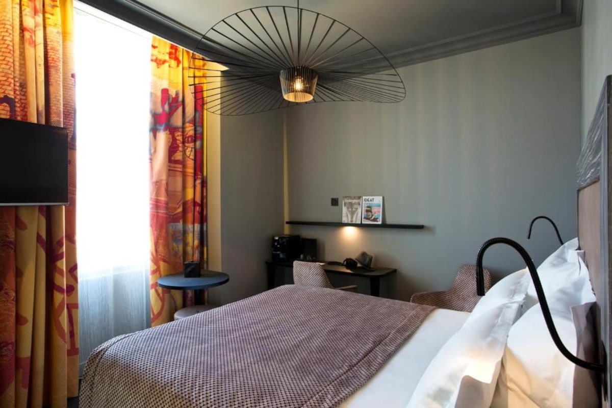Habitación de hotel con cama, escritorio, silla, televisor y una lámpara de techo original.