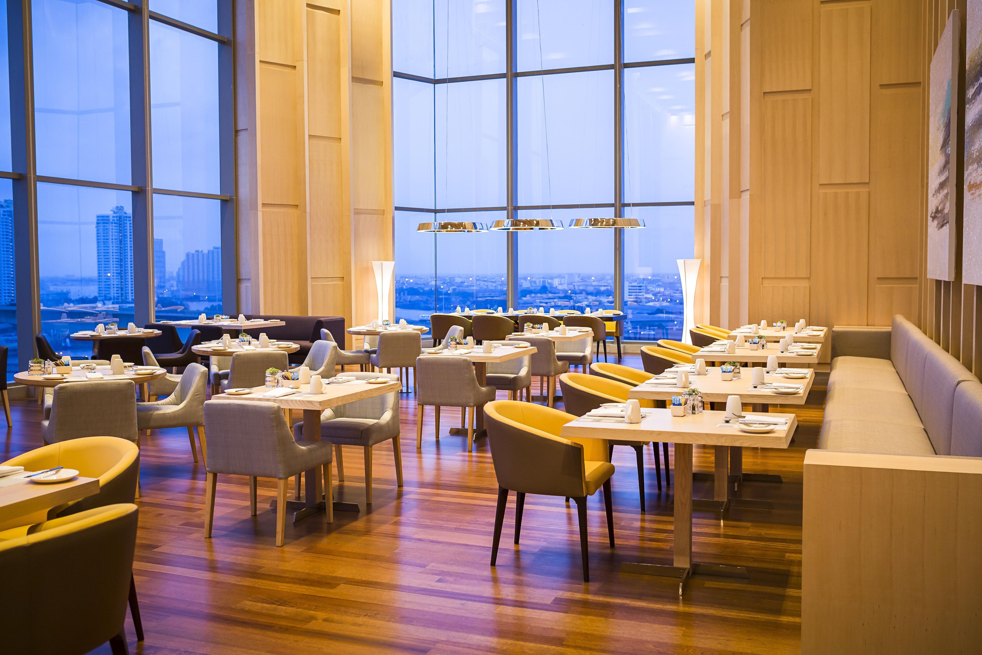 A modern restaurant with a city view, featuring round tables, chairs, and a wooden floor.