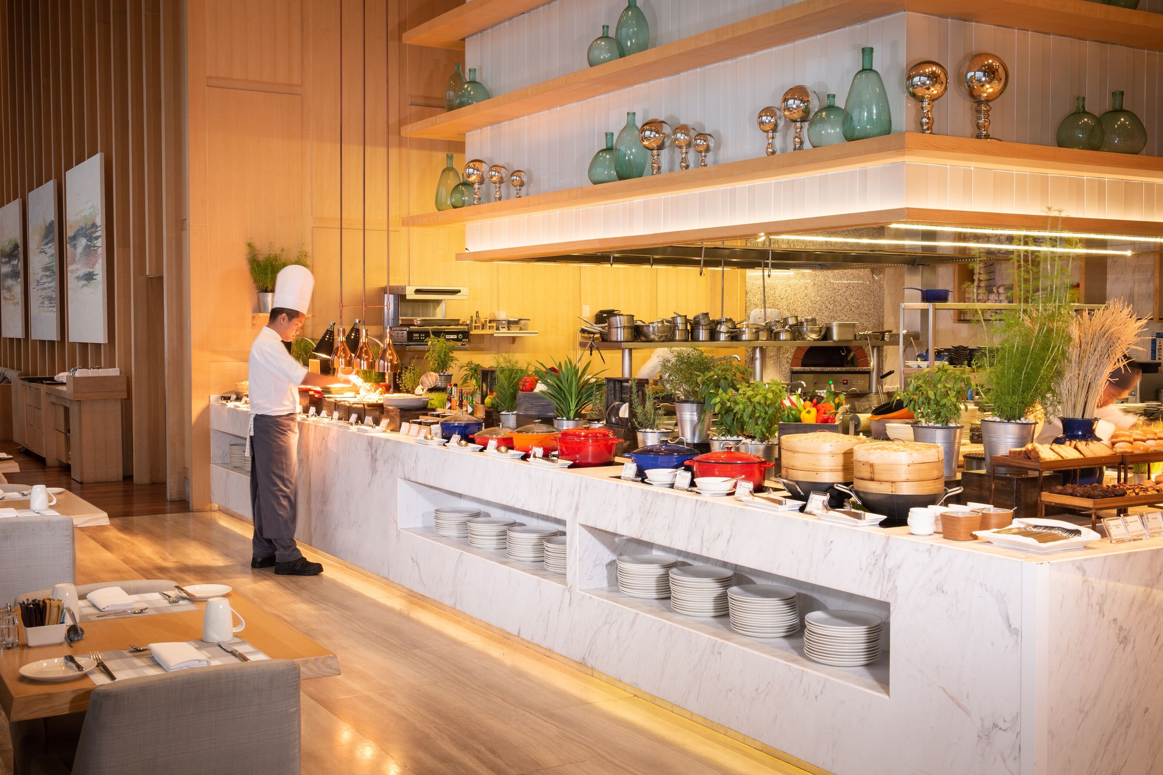 A modern kitchen with a chef preparing food, a buffet counter with various dishes, and a dining area with tables set for guests.