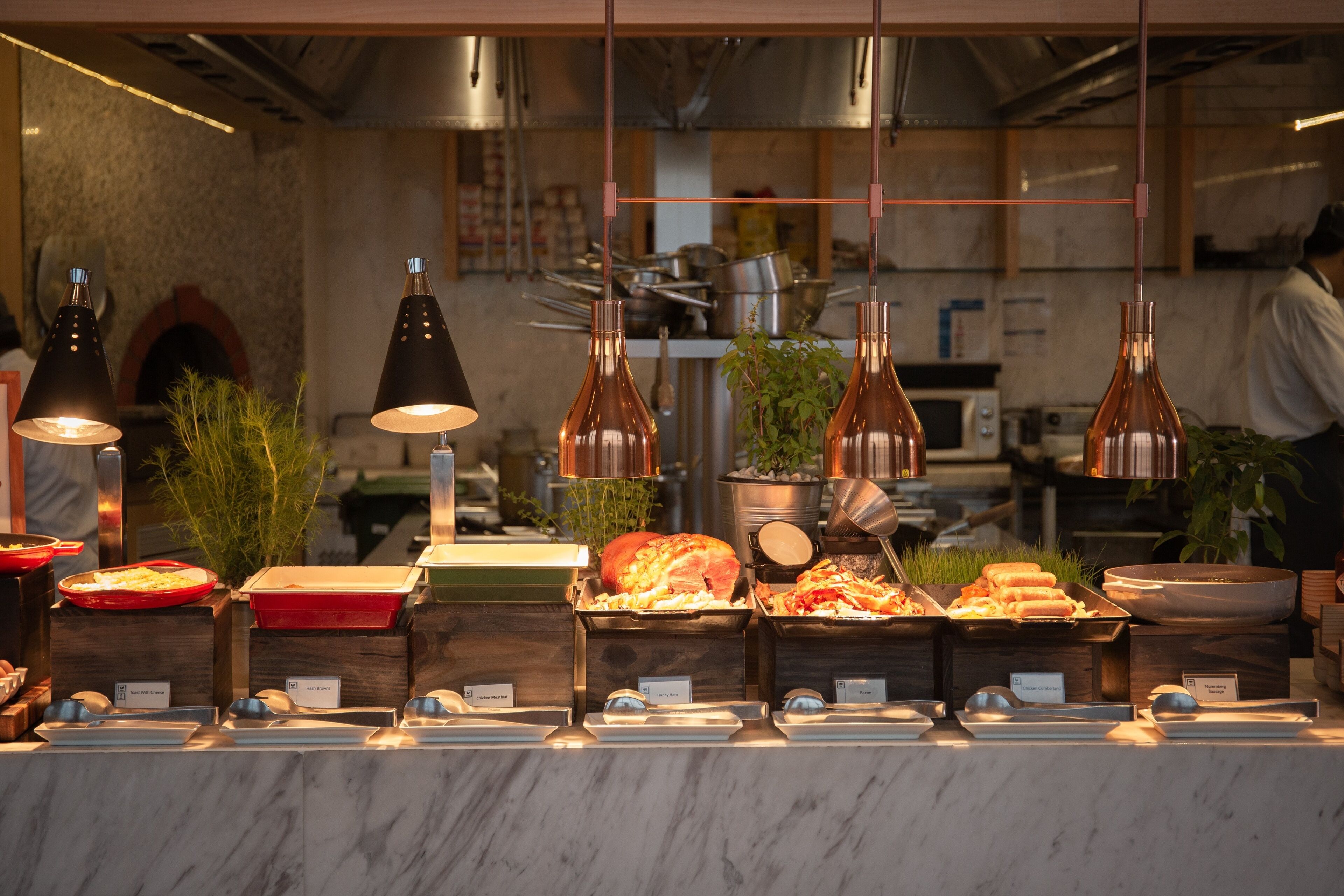 A buffet setup with various food items and serving utensils, set in a kitchen environment with visible appliances and a chef in the background.