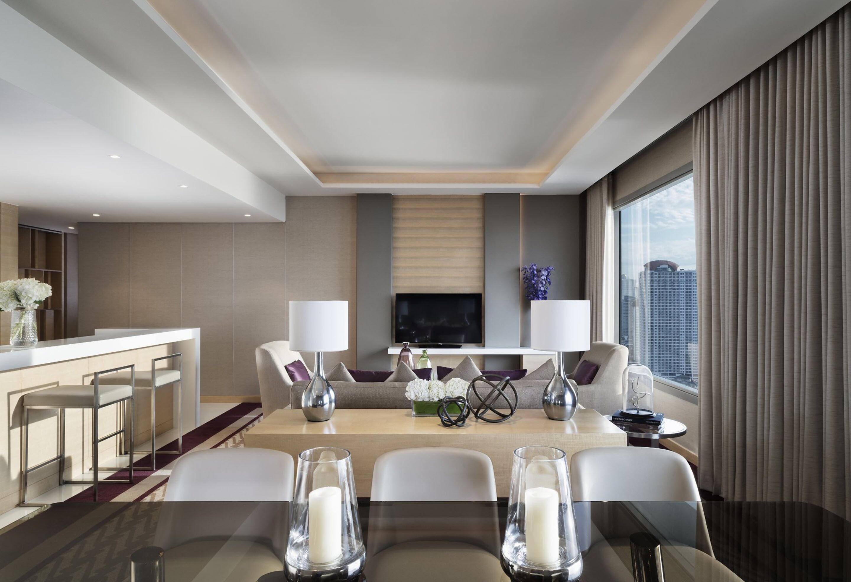Una habitación de hotel moderna con comedor, televisor montado en la pared y un ventanal con cortinas.