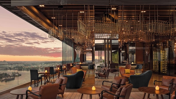A rooftop lounge with leather chairs and tables, a bar area, and a view of the city skyline at sunset. - Avani+ Riverside Bangkok Hotel (Bangkok)