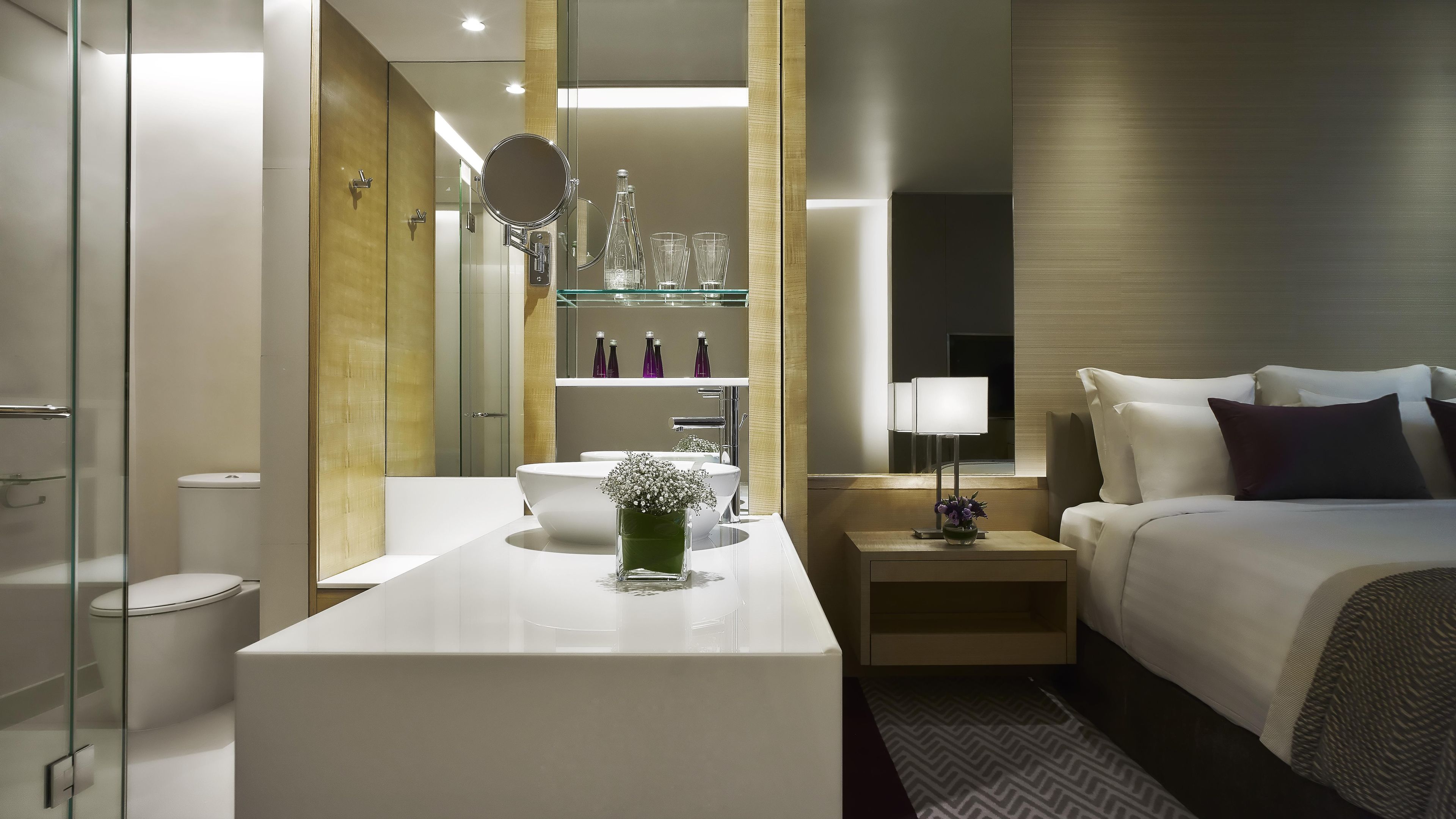 Une chambre d’hôtel moderne avec un grand lit, une table de chevet avec une lampe, une salle de bain équipée de toilettes, d’un lavabo et d’un miroir.