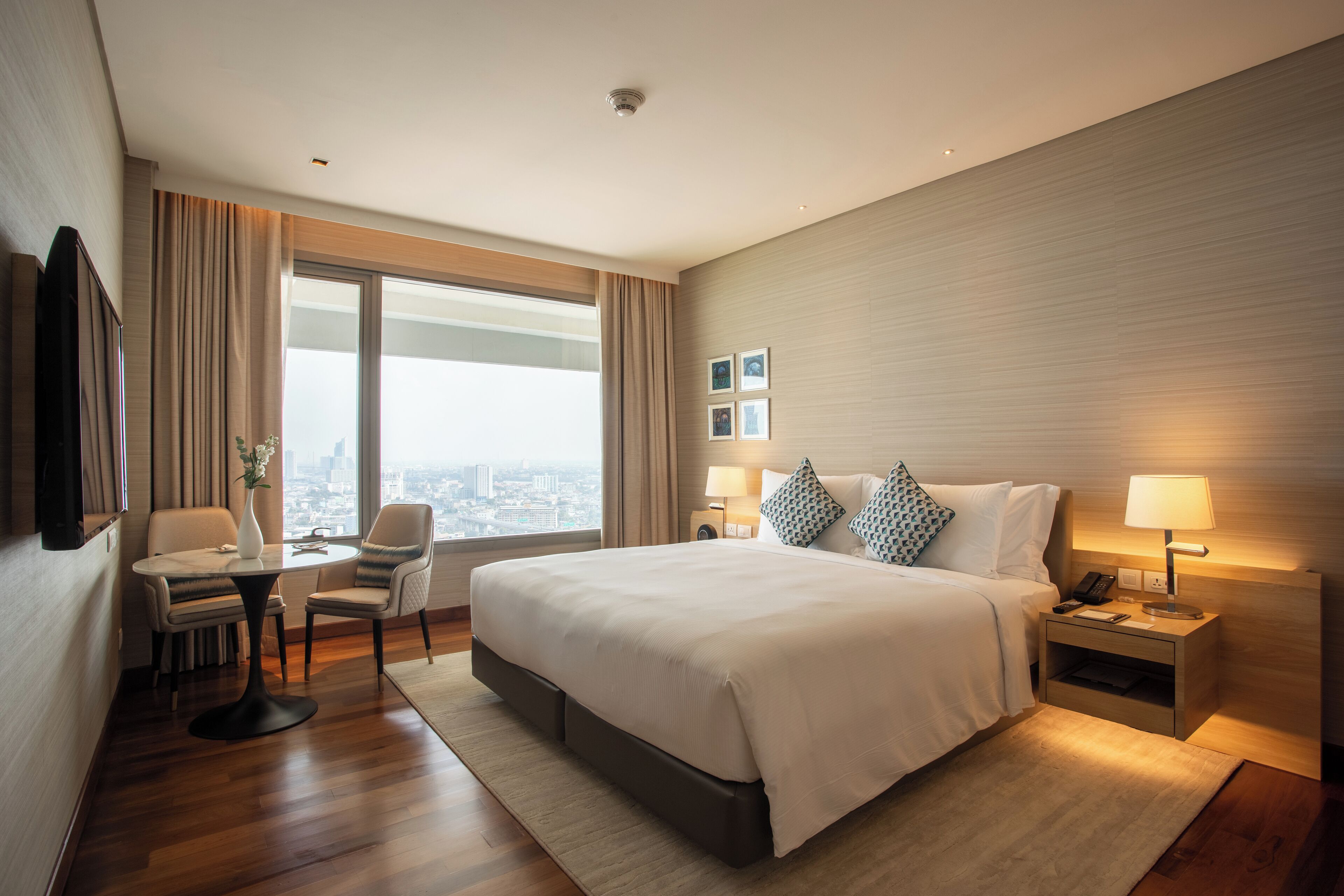 Quarto de hotel moderno com uma cama grande, uma escrivaninha com duas cadeiras, vista para a cidade e uma mesa de cabeceira com abajur.
