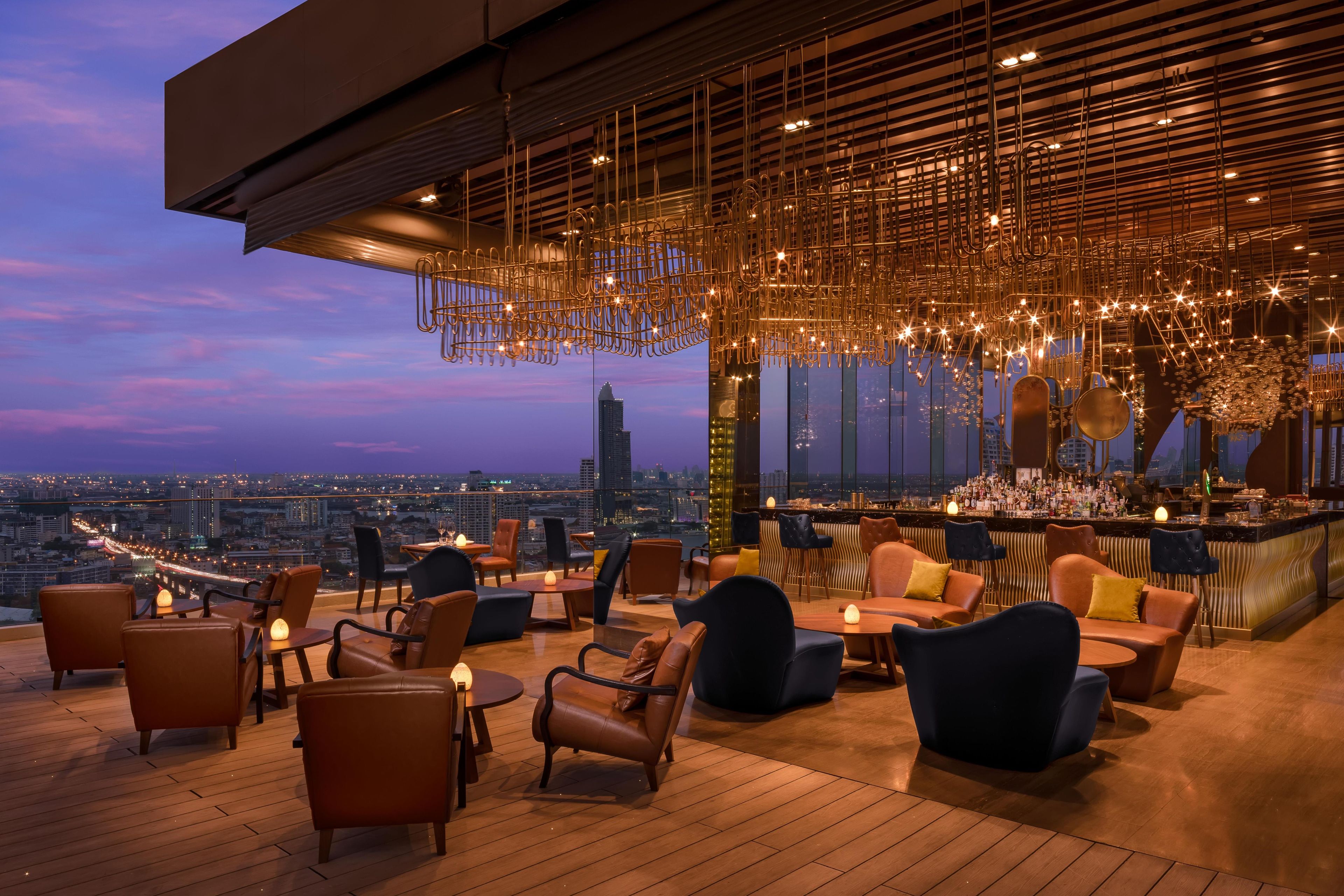 Un bar situé sur le toit d’un immeuble, offrant une vue sur la ville, doté d’un mobilier moderne et agrémenté d’un luminaire décoratif au plafond.