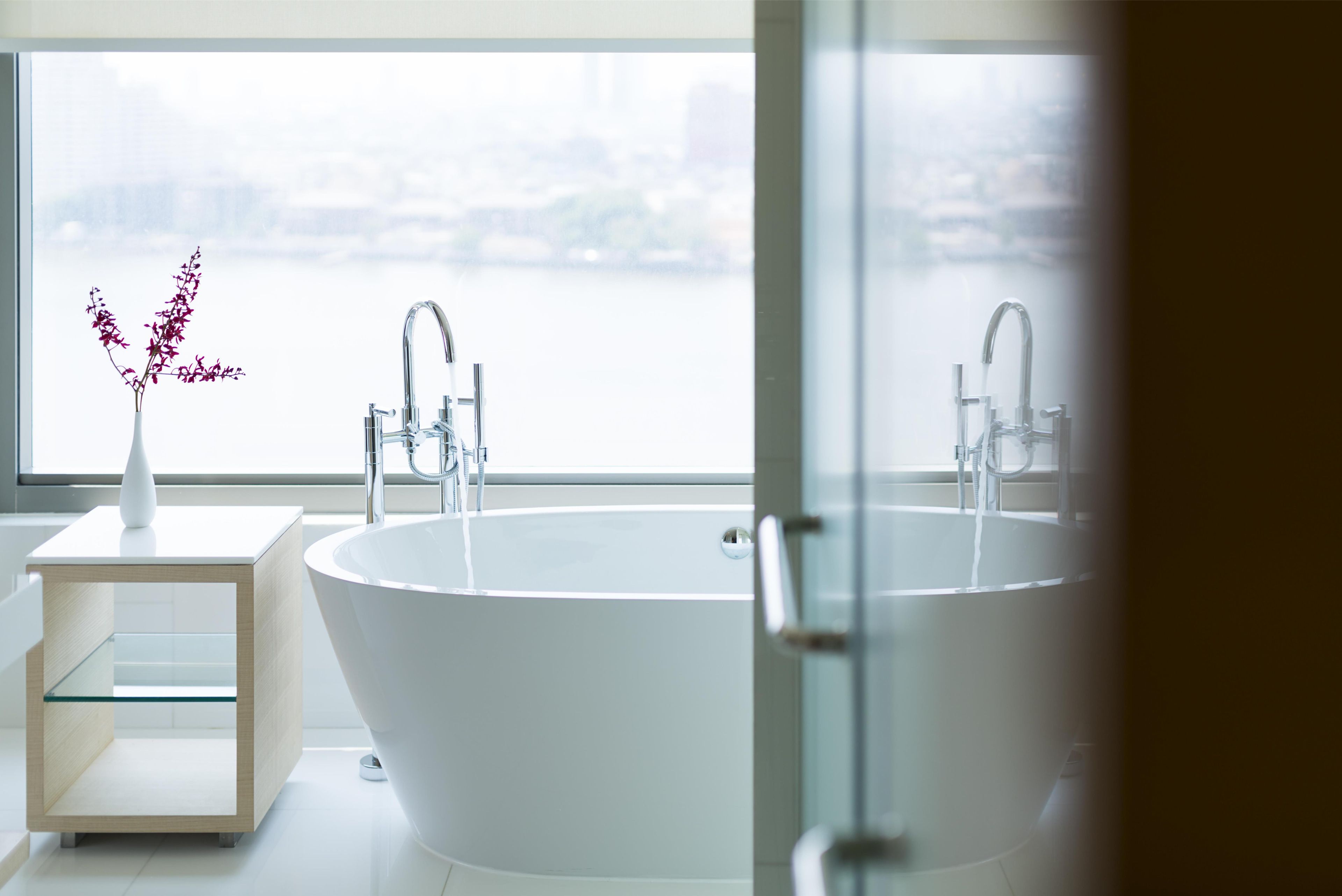 Un baño moderno con una bañera independiente, una mesita auxiliar con un florero y vistas a un paisaje urbano a través de un ventanal grande.