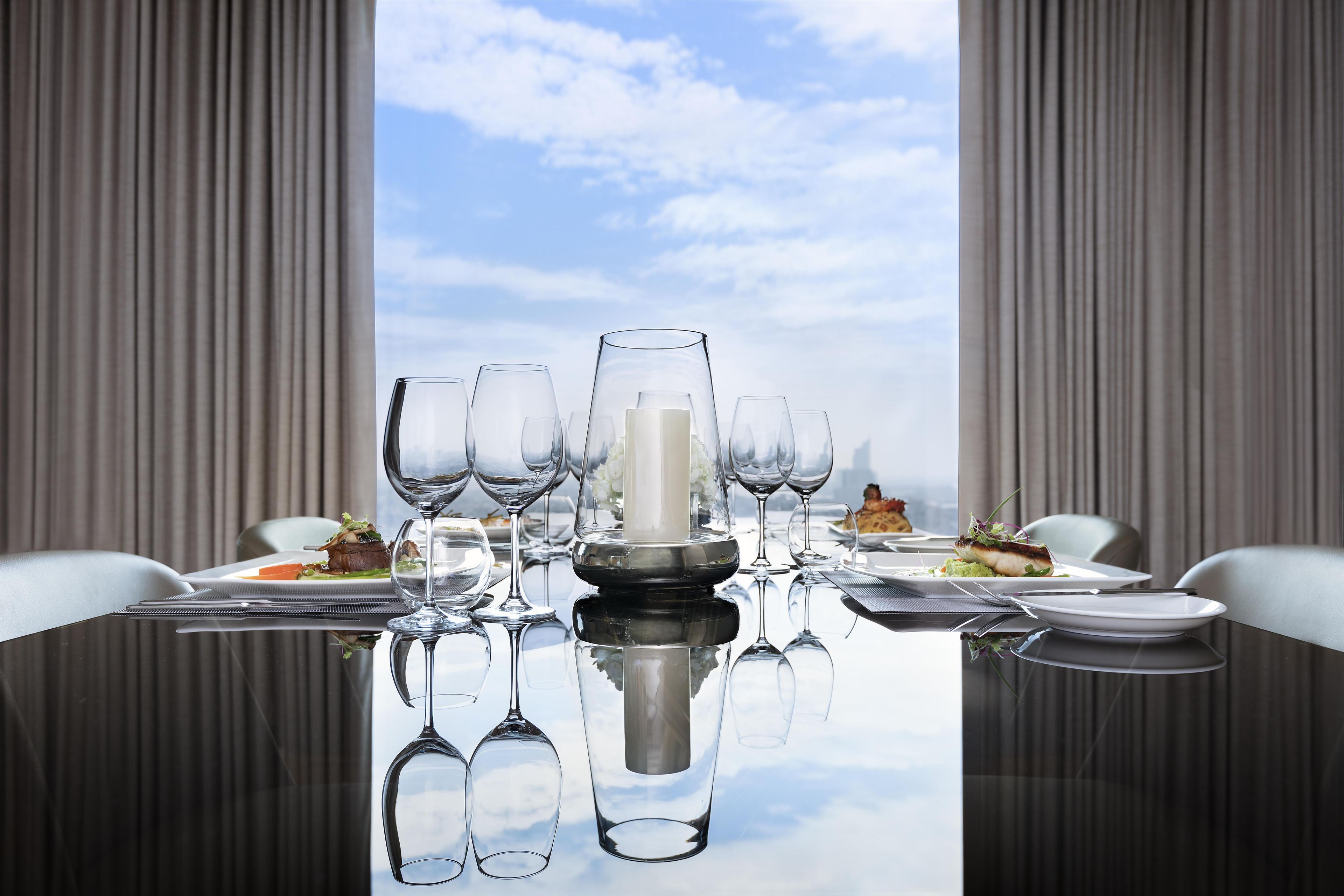 A dining table set with multiple wine glasses, plates, and a central decorative item, reflecting on a glass surface with a view of the sky through a window.