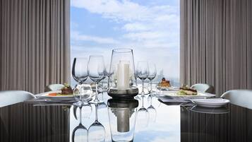 A dining table set with multiple wine glasses, plates, and a central decorative item, reflecting on a glass surface with a view of the sky through a window.