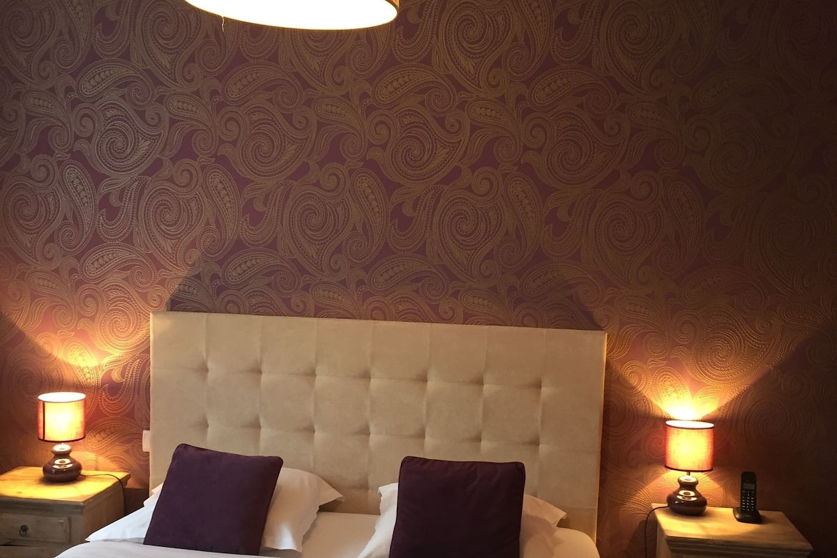A bedroom with a tufted headboard, two bedside lamps, and patterned wallpaper.