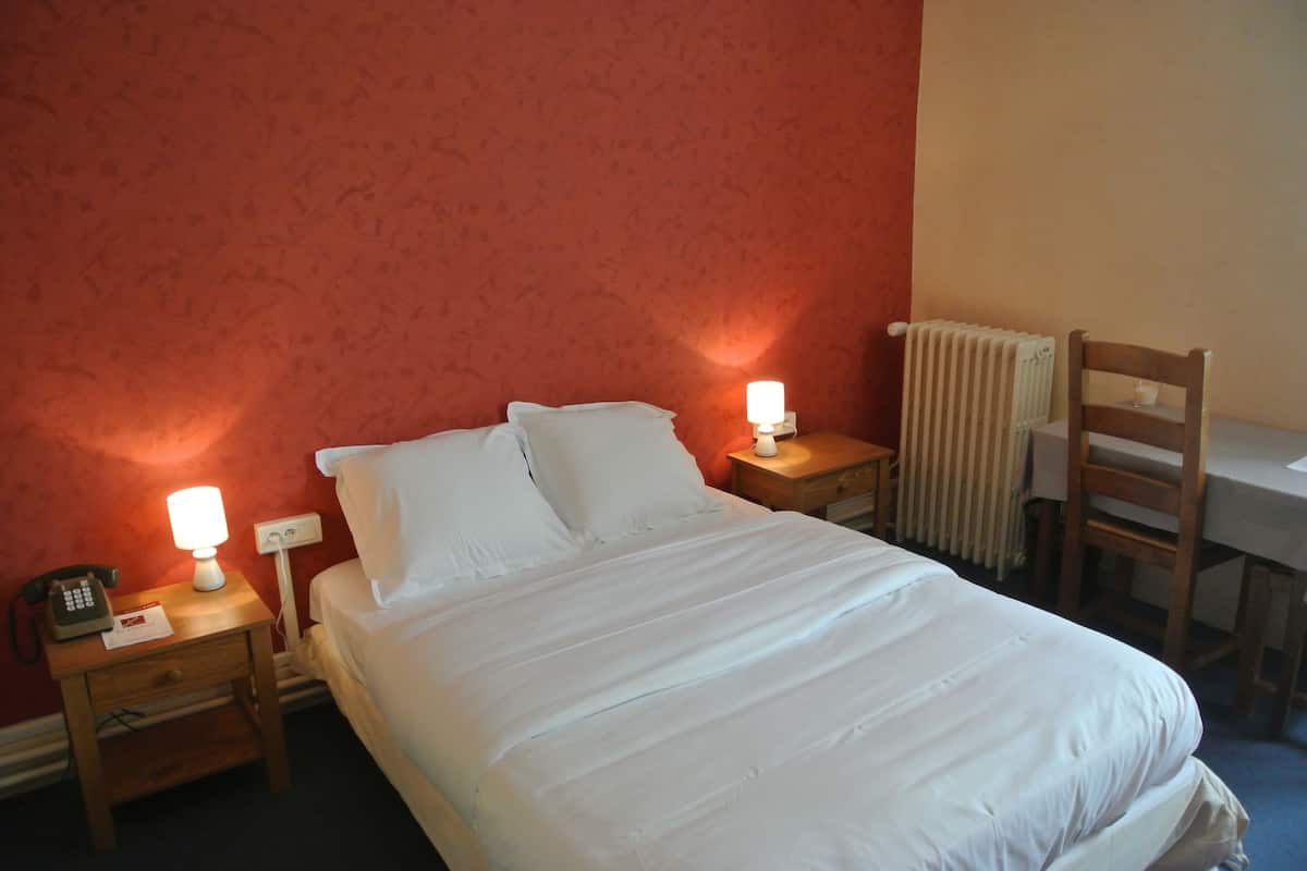 A neatly made bed with white linens, two bedside lamps, a wooden nightstand, a chair, and a radiator.