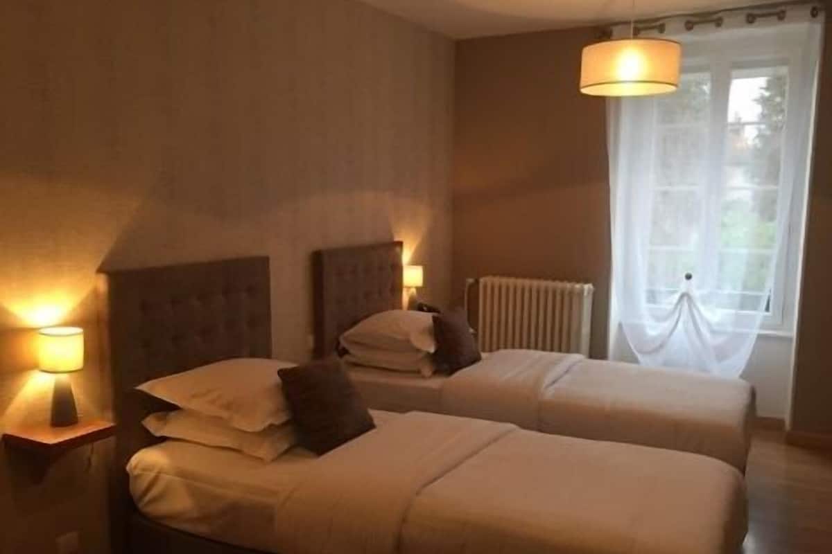 A hotel room with two beds, a radiator, a window with sheer curtains, and wall-mounted lamps.