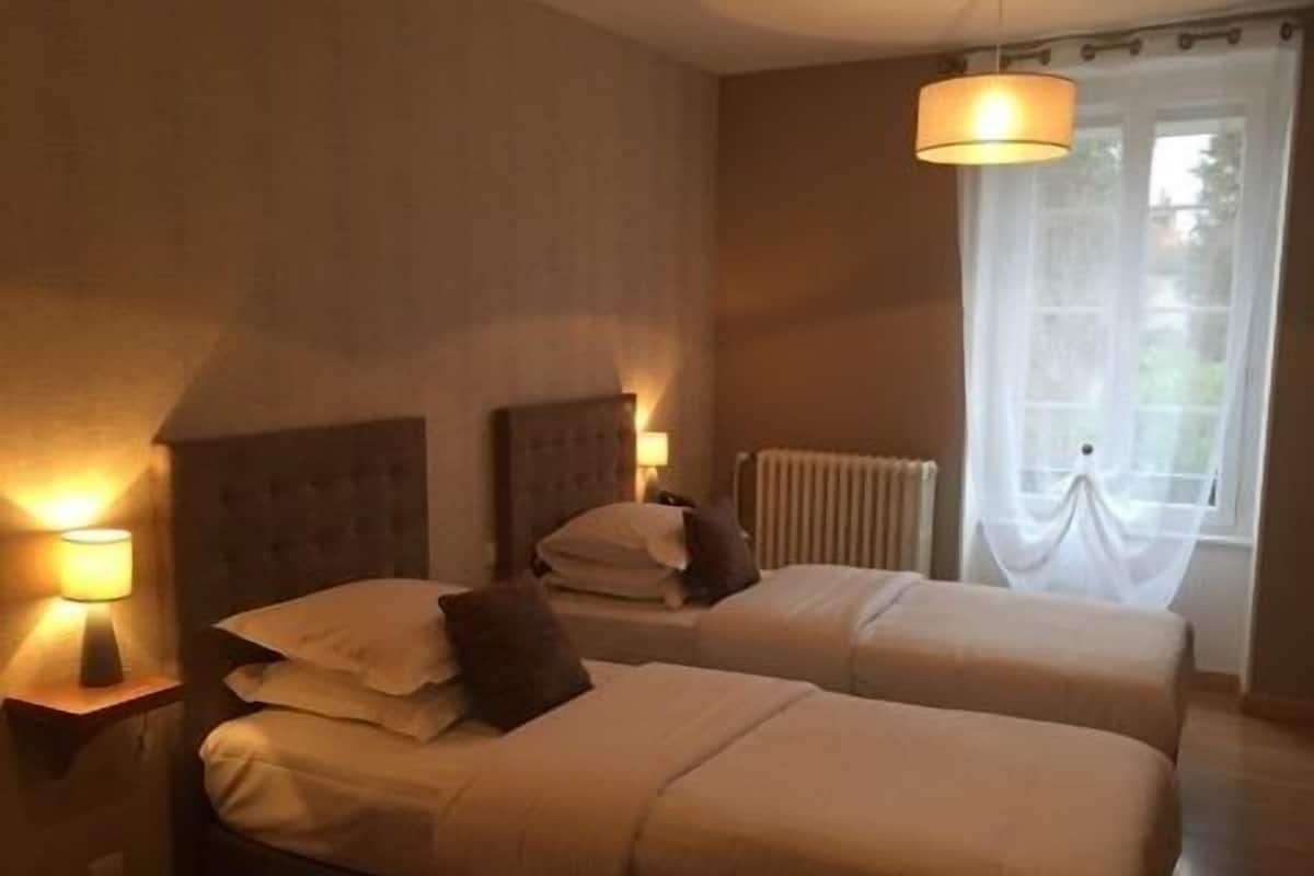 A hotel room with two beds, each with a headboard, a bedside table with a lamp, and a window with sheer curtains.