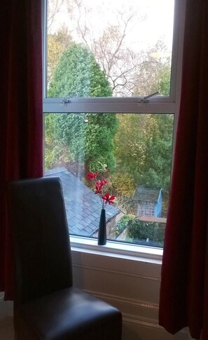 A room with a window, a chair, and a view of a garden with trees and a birdhouse. - Albany House (Penrith)