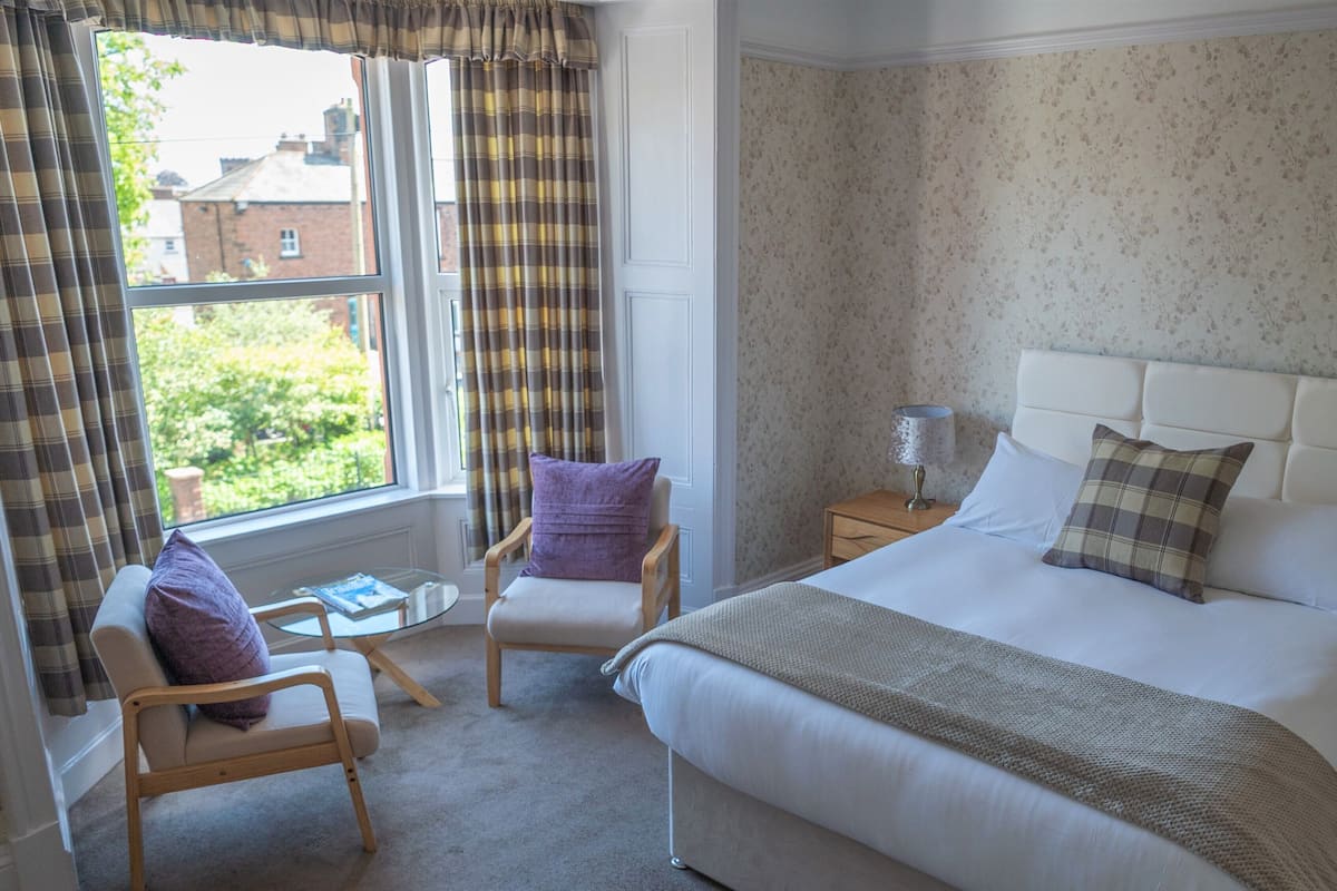 A hotel room with a bed, a chair, a small table, and a window with curtains.