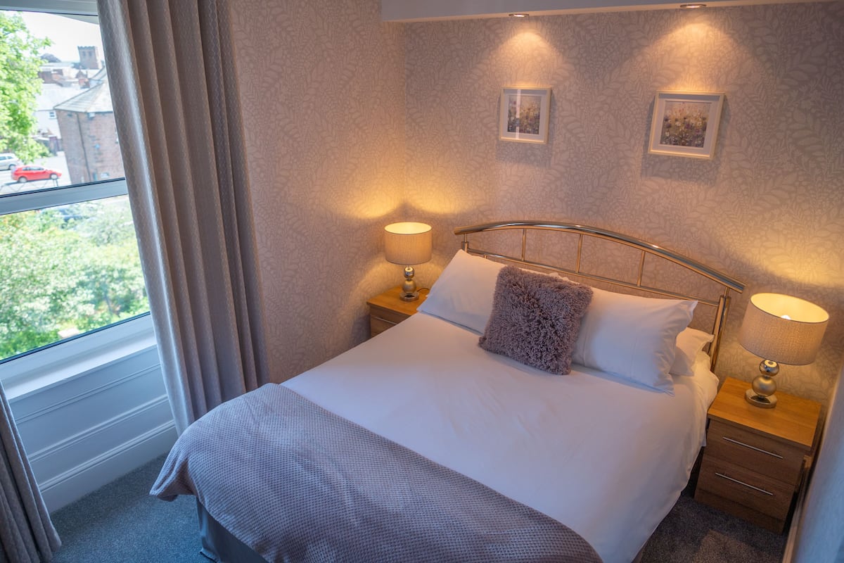 A neatly made bed with a headboard, two bedside lamps, and a window with curtains.