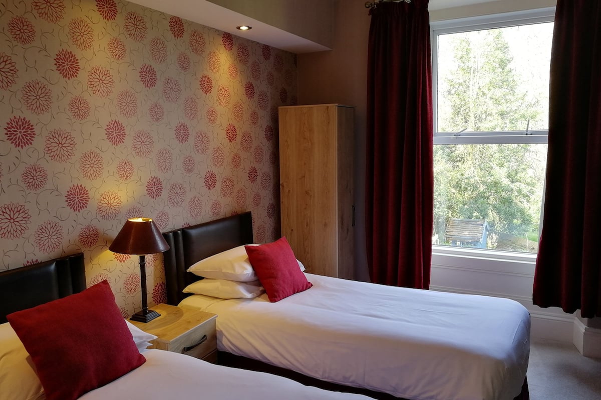 A hotel room with two beds, a nightstand, a lamp, a wardrobe, and a window with curtains.