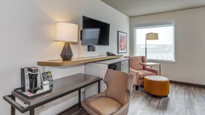 A modern hotel room with a flat-screen TV, a coffee maker, a lamp, a chair, a small table, and a window with a view of a snowy landscape.