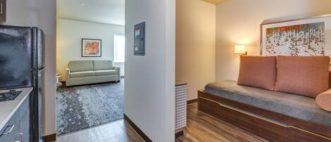 Una habitación de hotel moderna con kitchenette, un sofá gris con dos cojines, una alfombra estampada y un cuadro en la pared.