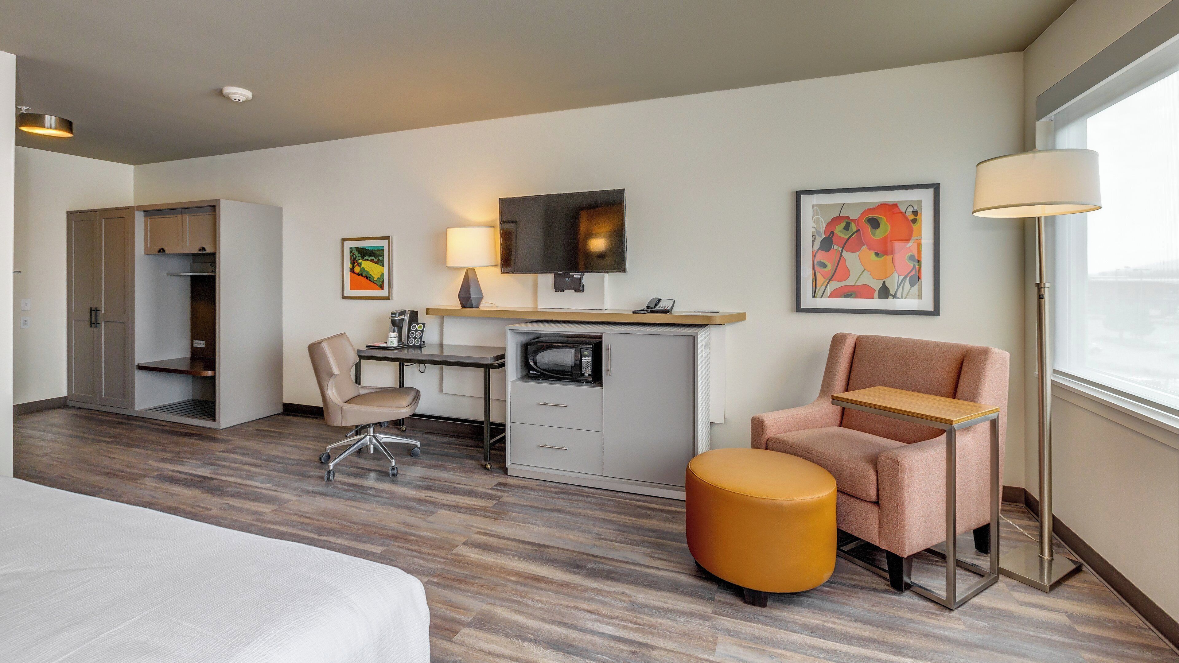 Een hotelkamer met een bed, een bureau met een computer, een stoel, een magnetron, een koffiezetapparaat, een televisie en kunstwerken aan de muren.
