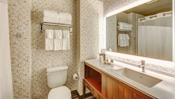 Un baño con inodoro, lavamanos y espejo. Toallas cuelgan de un perchero sobre el inodoro.
