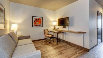 Habitación de hotel con sofá, escritorio con lámpara, una silla, televisor de pantalla plana y una puerta.