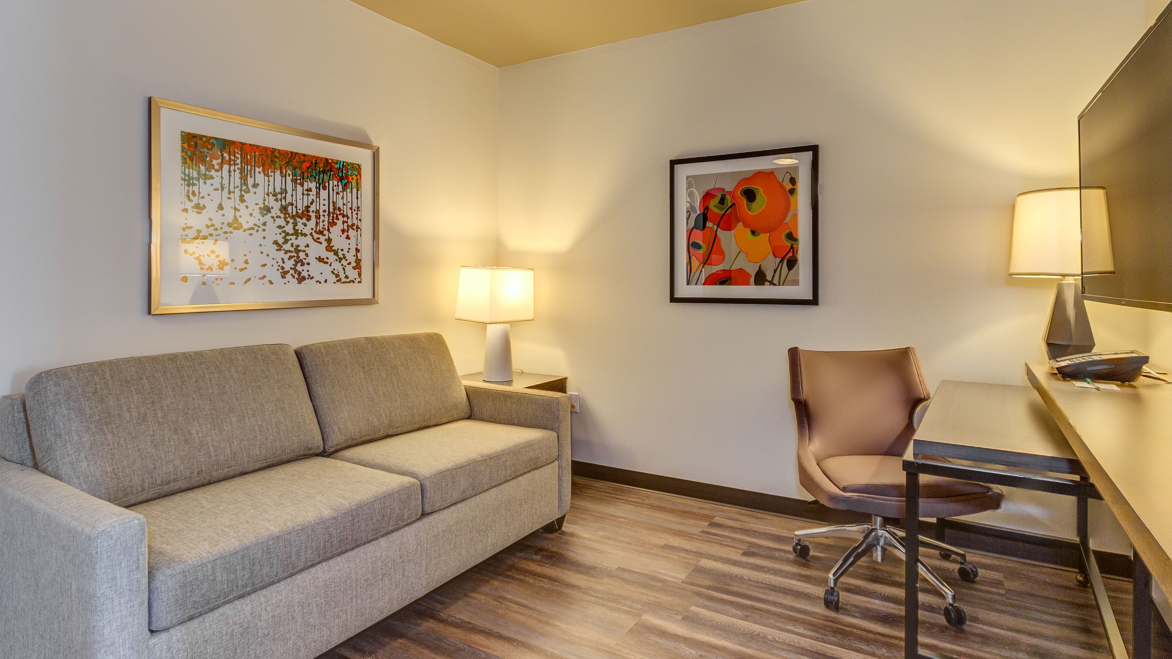 A hotel room with a sofa, a chair, a desk, and two lamps. There are two framed artworks on the walls.