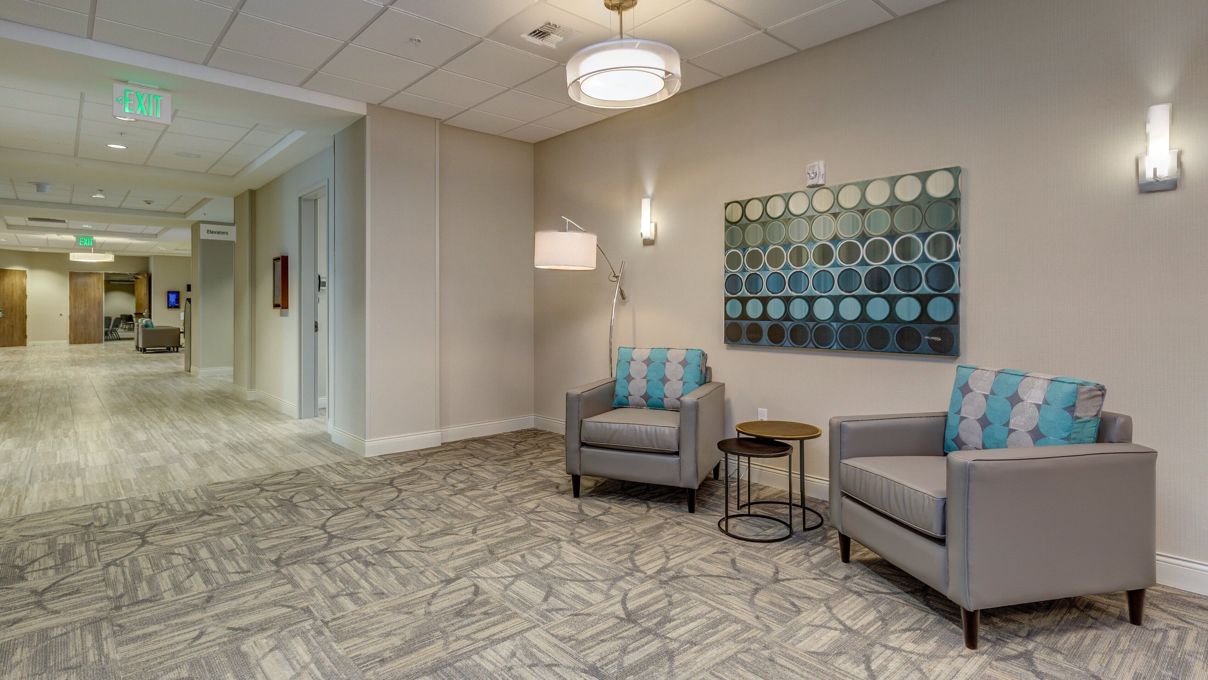A hotel lobby with a patterned carpet, two armchairs with patterned cushions, a floor lamp, and a wall art piece.