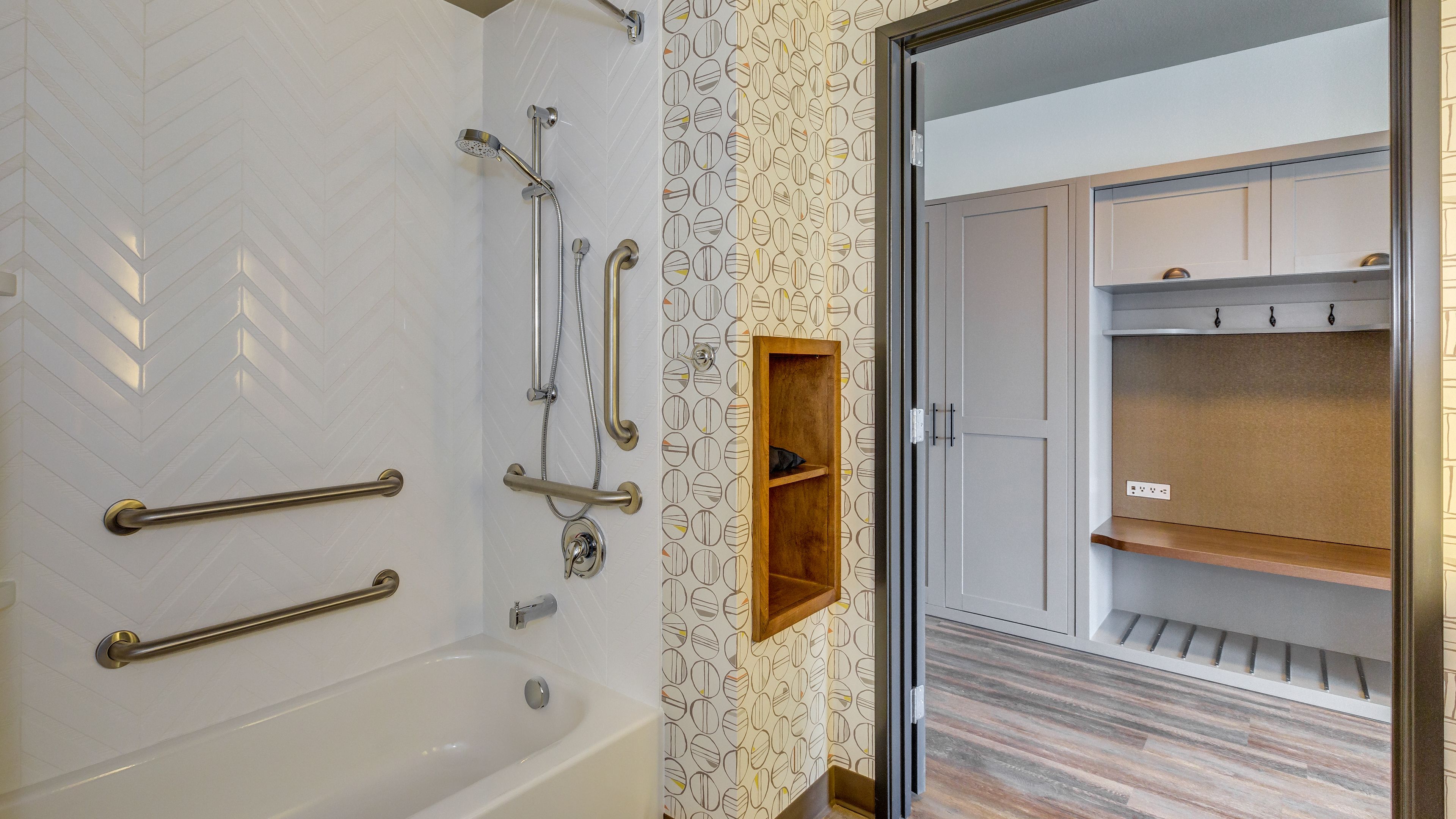 A modern bathroom with a bathtub, shower, and handrails. There is a wooden shelf and a wardrobe with a mirror and hooks.