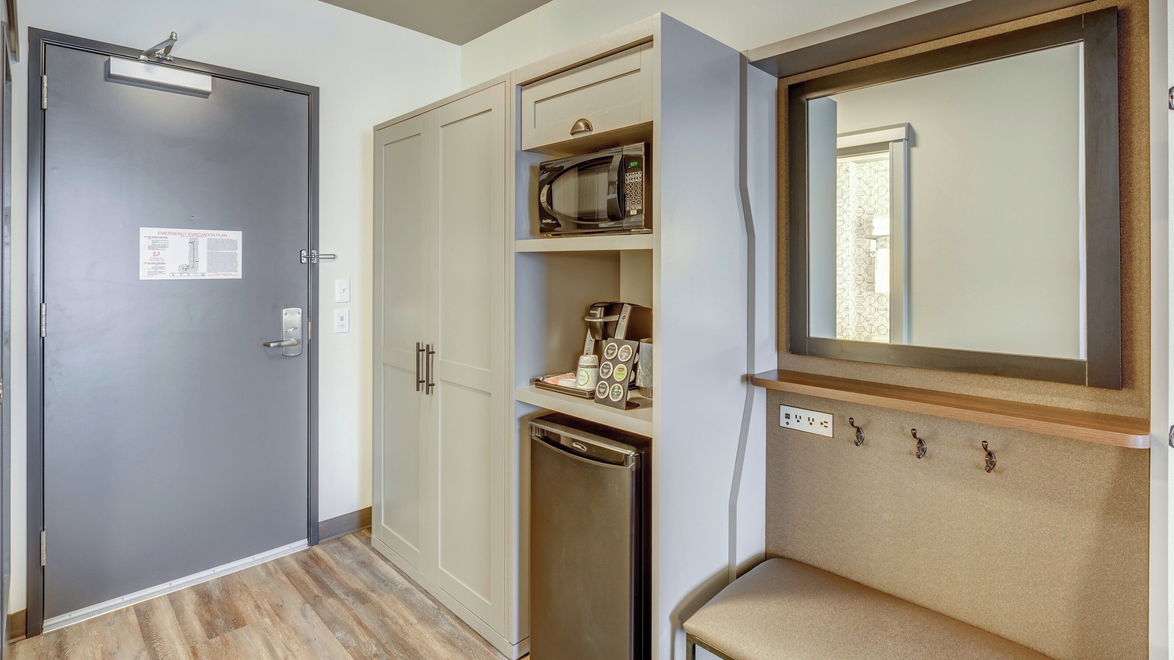 A hotel room with a grey door, a wardrobe, a microwave, a coffee maker, and a mirror.