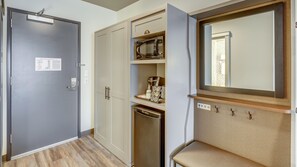 A compact hotel room with a grey door, a wardrobe, a microwave, a coffee maker, and a small seating area with a mirror.