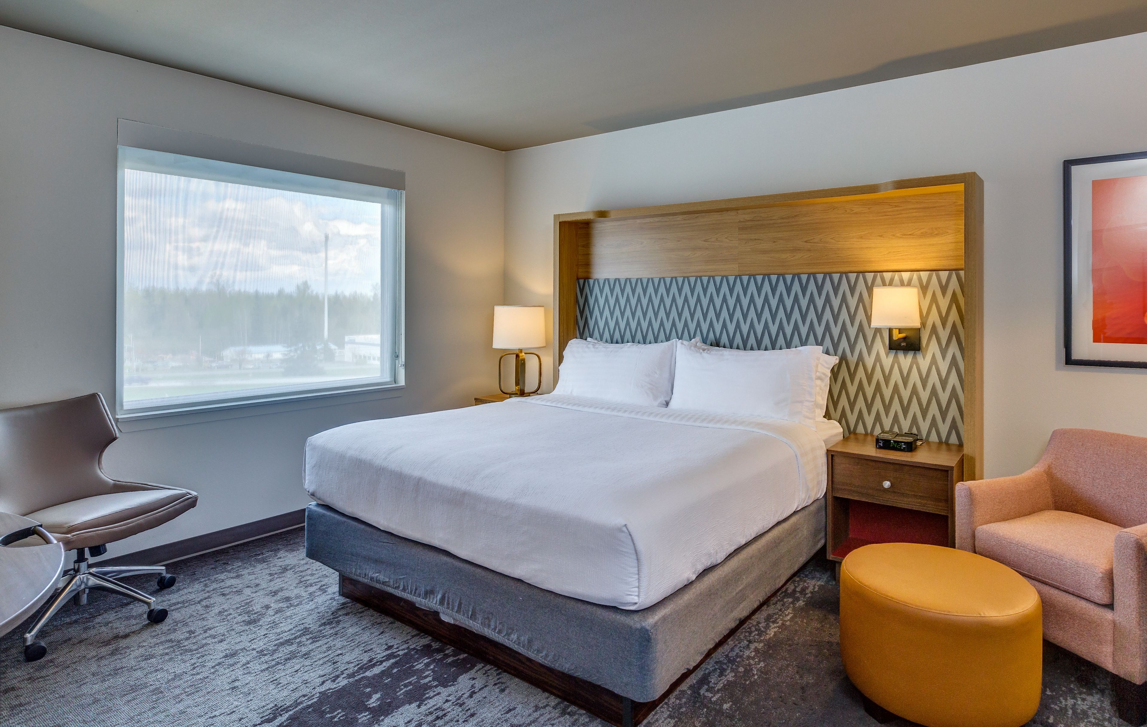 A hotel room with a large bed, two bedside tables with lamps, a chair, and a window with a view of trees.