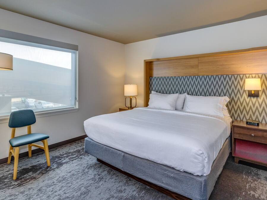 Habitación de hotel con una cama grande, una silla, un escritorio y un ventanal con persianas.