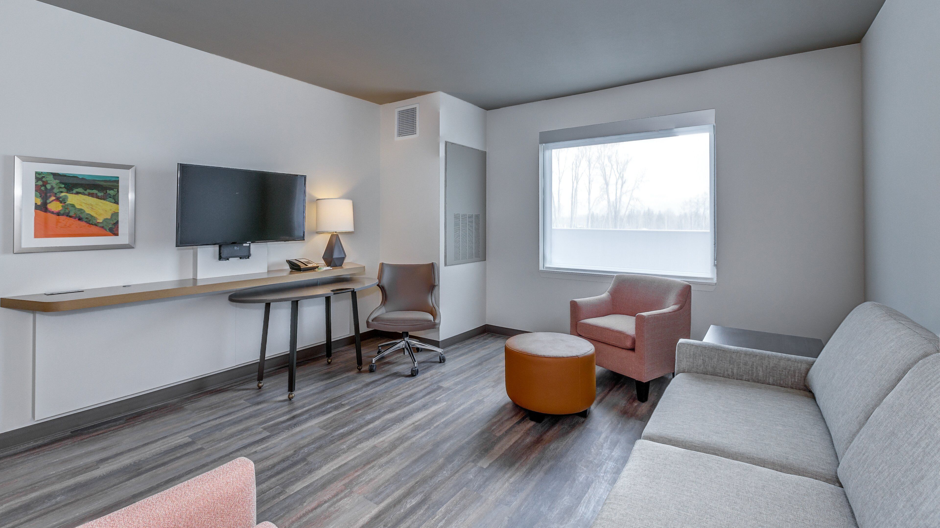 Moderni hotellihuone, jossa on taulutelevisio, työpöytä kahdella tuolilla, sohva ja suuri ikkuna.